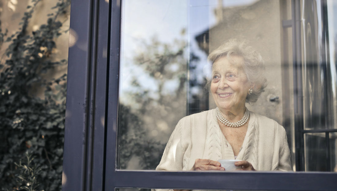 older woman stands in front of a window, smiling and holding a cup of tea or coffee