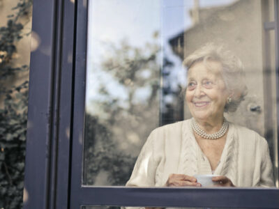 older woman stands in front of a window, smiling and holding a cup of tea or coffee