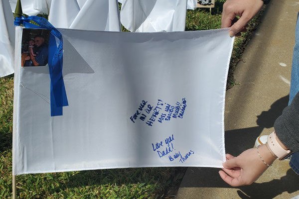 person extends a white flag with a photo of a man and a written message that says "Forever in our hearts! We miss you sooo much" and "Miss you Dada!" Baby Thomas