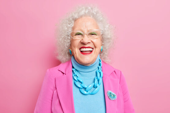Portrait of aged beautiful woman with curly grey hair bright makeup smiles happily expresses positive emotions dressed in fashionable outfit isolated over pink background. Positive grandmother