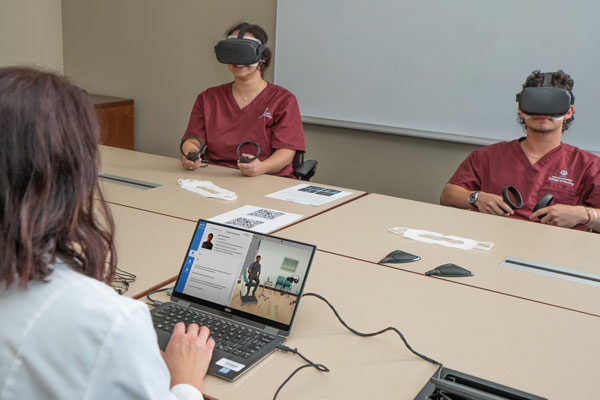 two nursing students wear virtual reality goggles and take part in a simulation run by a nursing professor who sits in front a laptop computer