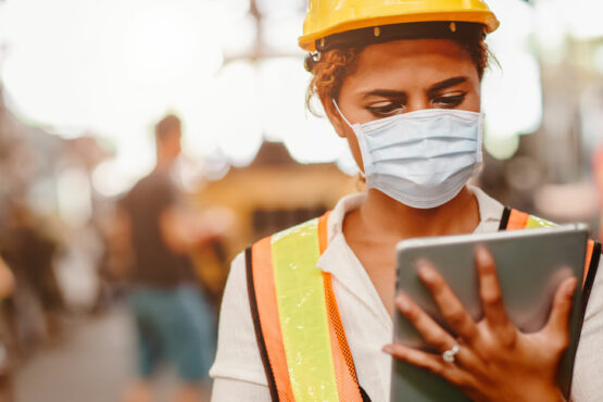 female worker wears a hard hat and face mask while looking at a tablet in an industrial setting